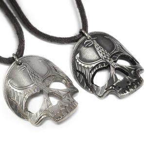 Op de afbeelding: Twee zilveren schedel hangers met ingewikkelde ontwerpen, hangend aan zwarte koorden. De schedels zijn uitgesneden en hebben een vogelontwerp erin.