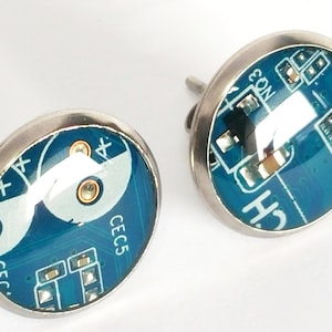 Pode incluir: Um par de brincos de circuito azul e branco com configurações prateadas. Os brincos apresentam um close-up de um circuito impresso com o texto "CEC5" e "CON" visível.