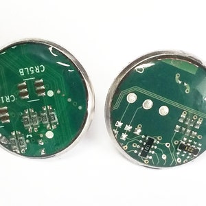 Könnte beinhalten: Ein Paar runde, silberne Ohrringe mit grünen Platinenmustern, die in klarem Harz eingebettet sind. Die Platinen haben den Text "CR1BU" und "CRS1B".