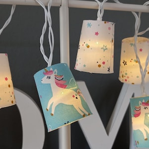 Puede incluir: Cadena de luces decorativas con pequeñas pantallas cónicas. Algunas pantallas presentan un diseño de unicornio sobre un fondo azul, otras un fondo blanco con estrellas y puntos de colores. Las luces están ensartadas en un cable blanco.