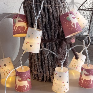 Op de afbeelding: Een string met lampjes met papieren lantaarns met een eenhoornontwerp. De lantaarns zijn in tinten roze, wit en crème met gouden glitteraccenten. De lampjes zijn aan een doorzichtig draad geregen en zijn verlicht met warmwitte lampjes.
