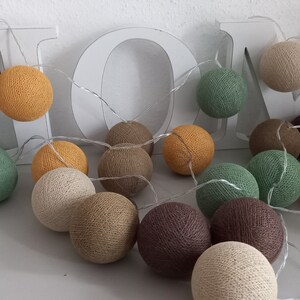 May include: A string of cotton ball lights in various shades of brown, beige, green, and yellow. The lights are strung together with a clear wire and are hanging from a white wooden sign that says "HOME".