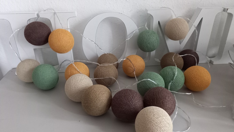 May include: A string of cotton ball lights in various shades of brown, beige, green, and yellow. The lights are strung together with a clear wire and are hanging from a white wooden sign that says "HOME".