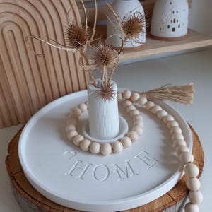 Può includere: Un vassoio decorativo bianco con la scritta "HOME" in rilievo, circondato da una ghirlanda di perline di legno. Un piccolo vaso bianco contiene fiori secchi. Il vassoio poggia su una fetta di legno, con un decoro ad arco in legno sullo sfondo.