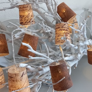 Op de afbeelding: Een lichtsnoer met bruine papieren lantaarns die om witte takken zijn gewikkeld. De lantaarns hebben een vintage uitstraling met handgeschreven tekst.