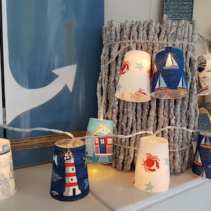 Può includere: Catena luminosa con paralumi a forma di bicchieri di carta a tema nautico. I paralumi presentano disegni di barche a vela, fari, granchi e case sulla spiaggia. Le luci sono infilate su un filo sottile ed emettono un bagliore caldo.