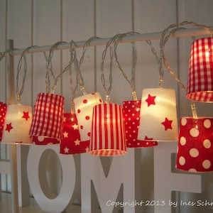 Può includere: Una catena di luci bianche con paralumi in tessuto rosso e bianco. I paralumi hanno diversi motivi, tra cui gingham, pois e strisce. Le luci sono appese a un'asta bianca e compongono la parola "HOME".