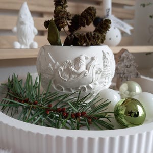Puede incluir: Una bandeja blanca acanalada contiene adornos navideños, ramitas de pino y una maceta decorativa con un diseño de trineo de Papá Noel. Una figura de gnomo blanco y otras decoraciones navideñas están en el fondo.