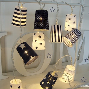 Peut inclure: Une guirlande lumineuse décorative avec de petits abat-jours cylindriques. Les abat-jours présentent divers motifs, notamment des rayures, des pois, des étoiles et du vichy, en noir et blanc. Les lumières sont suspendues à un fil fin, créant une ambiance fantaisiste et festive.