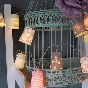 Könnte beinhalten: Eine Lichterkette mit Stofflampenschirmen in verschiedenen Mustern, darunter Streifen, Punkte und Blumenmuster. Die Lichter sind um einen dekorativen Vogelkäfig gewickelt.
