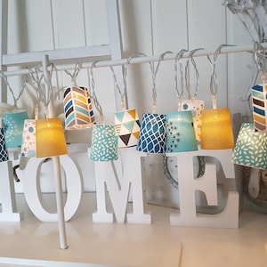 Può includere: Filo di luci con paralumi in tessuto in vari motivi geometrici di blu, giallo e marrone. Le luci sono appese a un'asta di metallo bianca e compongono la parola "HOME" in lettere bianche.