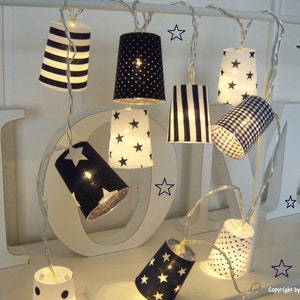 Può includere: Filo di luci con paralumi in carta bianca decorati con motivi in ​​bianco e nero, tra cui strisce, pois e stelle. Le luci sono infilate su un filo bianco e sono appese a una superficie bianca.