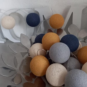 Könnte beinhalten: Eine Lichterkette aus Baumwollkugeln in verschiedenen Farben, darunter Beige, Grau, Blau und Gelb. Die Lichter sind in einer Schale mit einem Blattdesign angeordnet.