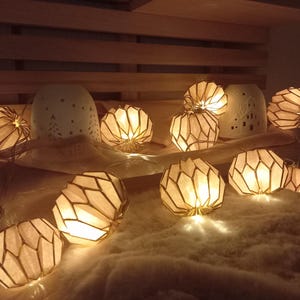 Op de afbeelding: Een warme, uitnodigende scène met verlichte papieren lantaarns met geometrische patronen. De lantaarns zijn geplaatst op een zacht, wit oppervlak en geven een zachte gloed. Op de achtergrond staan ​​verschillende keramische vazen.