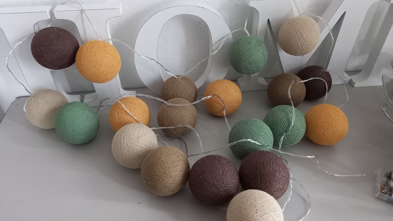 May include: A string of cotton ball lights with a variety of colors, including brown, beige, green, and yellow. The lights are strung together with a clear wire and are lit up.