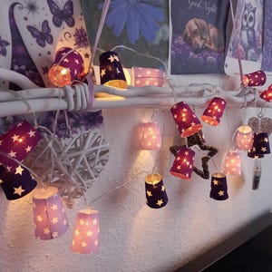 Può includere: Una stringa di luci decorative con ritagli a forma di stella. Le luci sono in tonalità viola e rosa, con un bagliore caldo. Sono disposte lungo un ramo bianco, creando un'atmosfera stravagante e festosa.
