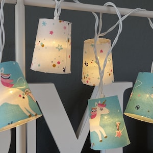 Può includere: Ghirlanda di luci con filo bianco e paralumi in tessuto blu e bianco. Ogni paralume presenta un design di unicorno con accenti rosa, oro e bianco. I paralumi sono decorati con stelle e coriandoli.