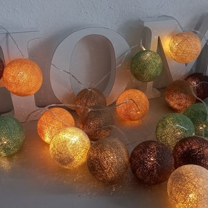 May include: A string of cotton ball lights with warm white LED bulbs. The lights are strung together with a clear wire and are arranged in a circle. The cotton balls are in various colors, including beige, brown, green, and orange.
