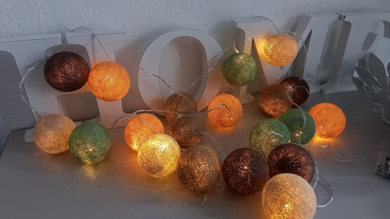May include: A string of cotton ball lights with warm white LED bulbs. The lights are strung together with a clear wire and are arranged in a circle. The cotton balls are in various colors, including beige, brown, green, and orange.