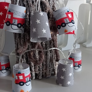 Può includere: Una ghirlanda di luci con paralumi in tessuto rosso e grigio. I paralumi presentano un design di camion dei pompieri rosso con stelle bianche su sfondo grigio. Le luci sono collegate da un filo trasparente.