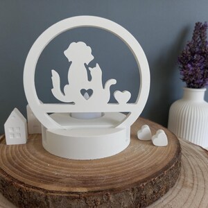 Könnte beinhalten: Weißer Kerzenhalter aus Holz mit einer Silhouette eines Hundes und einer Katze in einem Kreis. Der Kerzenhalter steht auf einem Holzsockel und es befinden sich zwei kleine weiße Herzen auf dem Sockel.