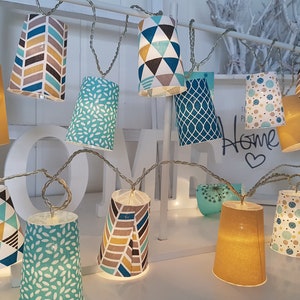 Pode incluir: Fio de luzes com abajures de papel estampados em várias cores, incluindo azul-petróleo, amarelo e castanho. Os abajures têm desenhos geométricos e florais. As luzes estão presas em um fio prateado.