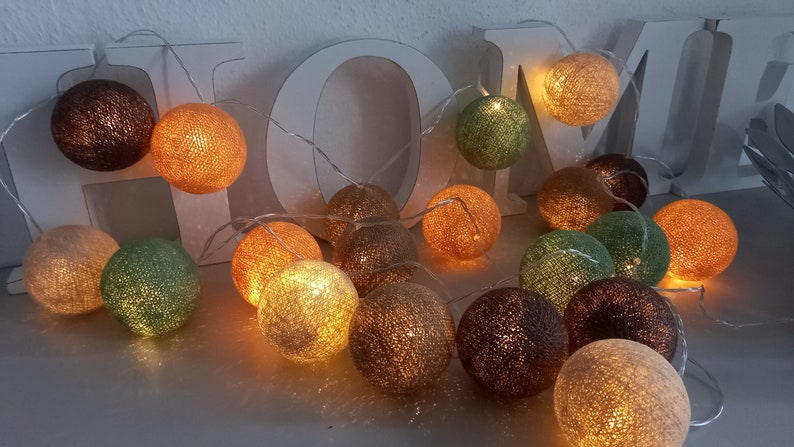 May include: A string of cotton ball lights with warm white LED bulbs. The lights are in various colors, including brown, orange, green, and beige. The lights are strung together with a clear wire and are hanging from a white wooden sign that says "HOME".