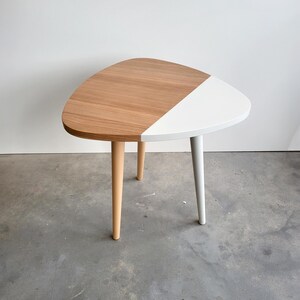 Könnte beinhalten: Ein dreieckiger Beistelltisch mit einer weißen und hölzernen Tischplatte und drei Holzbeinen. Der Tisch ist zur Hälfte weiß und zur Hälfte mit Holz lackiert.