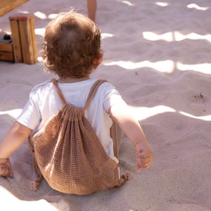Könnte beinhalten: Ein kleines Kind sitzt auf dem Sand und trägt ein weißes Hemd und eine braune Netz-Kordeltasche.