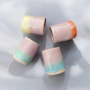 Könnte beinhalten: Vier Keramikbecher mit rosa Untergrund und einem Farbverlauf nach oben. Die Becher haben einen gelben, orangen, grünen und blauen Farbverlauf.
