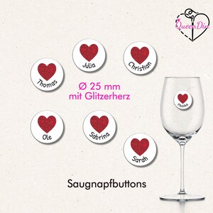 Könnte beinhalten: Weiße runde Weinglas-Anhänger mit roten Glitzerherz-Designs und Namen. Die Anhänger haben einen Durchmesser von 25 mm. Ein Anhänger ist an einem Weinglas befestigt. Der Text "Saugnapfbuttons" steht unten.