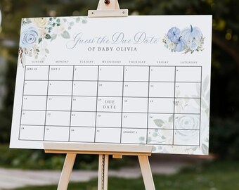 May include: A white sign on a wooden easel with the text "Guess the Due Date of Baby Olivia." The sign has a calendar grid with floral accents in blue and green. The background includes white balloons and greenery.