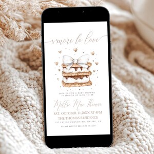 May include: A smartphone displays a baby shower invitation with the text "S'more to Love." The invitation features a watercolor illustration of a s'more with a bow and hearts. The phone rests on a cream-colored knitted blanket.