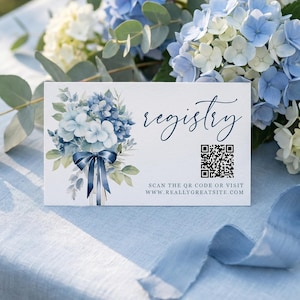 Könnte beinhalten: Eine weiße Registrierungskarte mit einem Aquarell-Blumenmuster aus blauen Hortensien und einem marineblauen Band. Das Wort "registry" ist in eleganter Schrift geschrieben. Ein QR-Code und eine Website-Adresse sind ebenfalls enthalten.