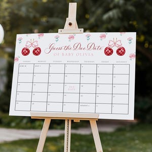 May include: A white sign on a wooden easel with the text "Guess the Due Date of Baby Olivia." The sign has a calendar grid with decorative red and pink accents, perfect for a baby shower game. The background is a blurred outdoor setting.