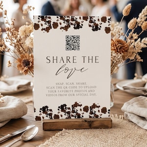 May include: A rectangular white sign with dark brown floral accents at the top and bottom. It features a black QR code, the text "SHARE THE love", and instructions: "SNAP. SCAN. SHARE. SCAN THE QR CODE TO UPLOAD YOUR FAVORITE PHOTOS AND VIDEOS FROM OUR SPECIAL DAY."