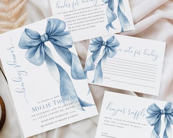 May include: Baby shower invitation set featuring a watercolor blue bow design. The set includes an invitation, a note card, a raffle card, and a card for guests to write a note. The cards are arranged on a white fabric background.
