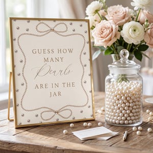 Op de afbeelding: Een goudkleurig ingelijst bord met de tekst GUESS HOW MANY Pearls ARE IN THE JAR, versierd met een parelrand en strikmotieven, geplaatst naast een doorzichtige glazen pot gevuld met witte parels en een boeket lichtroze en witte bloemen.