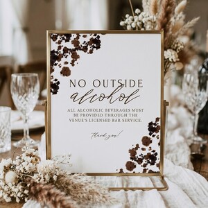 May include: A rectangular sign in a gold metal frame featuring white paper with dark brown watercolor-style splotches in the corners. The text reads: NO OUTSIDE alcohol, ALL ALCOHOLIC BEVERAGES MUST BE PROVIDED THROUGH THE VENUE'S LICENSED BAR SERVICE. Thank you!