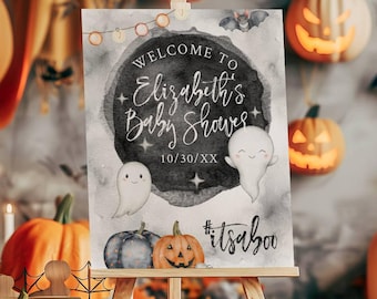 Puede incluir: Un letrero rectangular sobre un caballete de madera con un fondo gris estilo acuarela, dos fantasmas blancos, un centro circular oscuro y el texto Welcome to Elizabeth's Baby Shower, 10/30/XX, y #itsaboo, decorado con pequeñas estrellas.