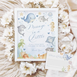 May include: A framed baby shower sign with the text "Message in a Bottle" and illustrations of marine life, including a whale, dolphins, and seahorses. A matching note card is included. The sign is surrounded by white flowers.