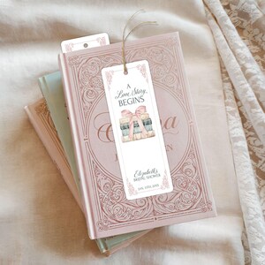 May include: A white bookmark with a pink ribbon and a stack of books. The bookmark says "A Love Story Begins" and "Elizabeth's Bridal Shower Jan. 12th, 20XX".