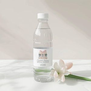 Puede incluir: Una botella de agua de plástico transparente con una etiqueta blanca. La etiqueta tiene una ilustración de acuarela de una pila de libros con una cinta rosa. El texto en la etiqueta dice "Elizabeth Anderson's Bridal Shower 4/12/20XX".