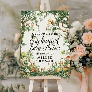 May include: A rectangular sign featuring a forest illustration with trees, vines, butterflies, and mushrooms. The text reads: Welcome to an Enchanted Baby Shower in honor of Millie Thomas. The sign is displayed on a light-colored wooden easel.