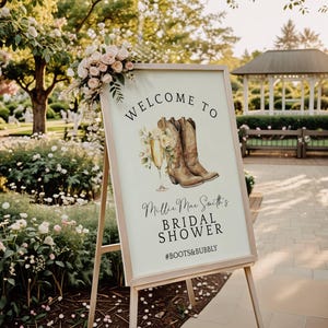 Peut inclure: Une pancarte de bienvenue pour une fête de mariage avec un thème rustique. La pancarte est en bois et présente une illustration aquarelle de bottes de cowboy et d'une flûte à champagne. Le texte sur la pancarte indique "Bienvenue à Millie Mae Smith's Bridal Shower #Boots&Bubbly".