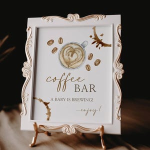 Pode incluir: Um sinal branco emoldurado com detalhes dourados, apresentando uma ilustração em aquarela de uma xícara de café com o texto "coffee bar" e "a baby is brewing! -enjoy!"