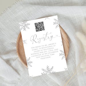 Puede incluir: Una tarjeta blanca con un diseño de copo de nieve plateado y el texto "Registry" en una fuente cursiva. La tarjeta también incluye un código QR y el texto "The bride-to-be, [bride's name], is registered at [name of boutique]. Wishing her joy, love, and laughter as we celebrate this exciting time together!"