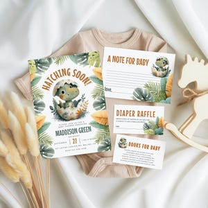 May include: A baby shower invitation set with a dinosaur theme. The invitation reads "Hatching Soon!" and includes details for Maddison Green's baby shower. The set includes cards for notes, diaper raffle, and books, along with a beige onesie and a wooden rocking horse.