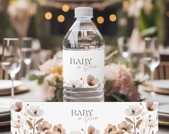 Op de afbeelding: Waterfleslabel met een bloemenontwerp en de tekst "Baby in Bloom". Het label is wit met bruine en beige bloemen en bladeren. Het label is ook verkrijgbaar als tafelkleed.