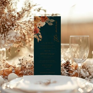 May include: A dark teal wedding menu card with floral accents in orange and white. The menu includes starters, main course, and dessert options. A wine glass, plate, and pinecone are also visible.